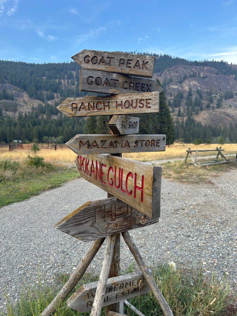 Wooden directional signs in Mazama, Washington, pointing toward local shops and hiking trails.